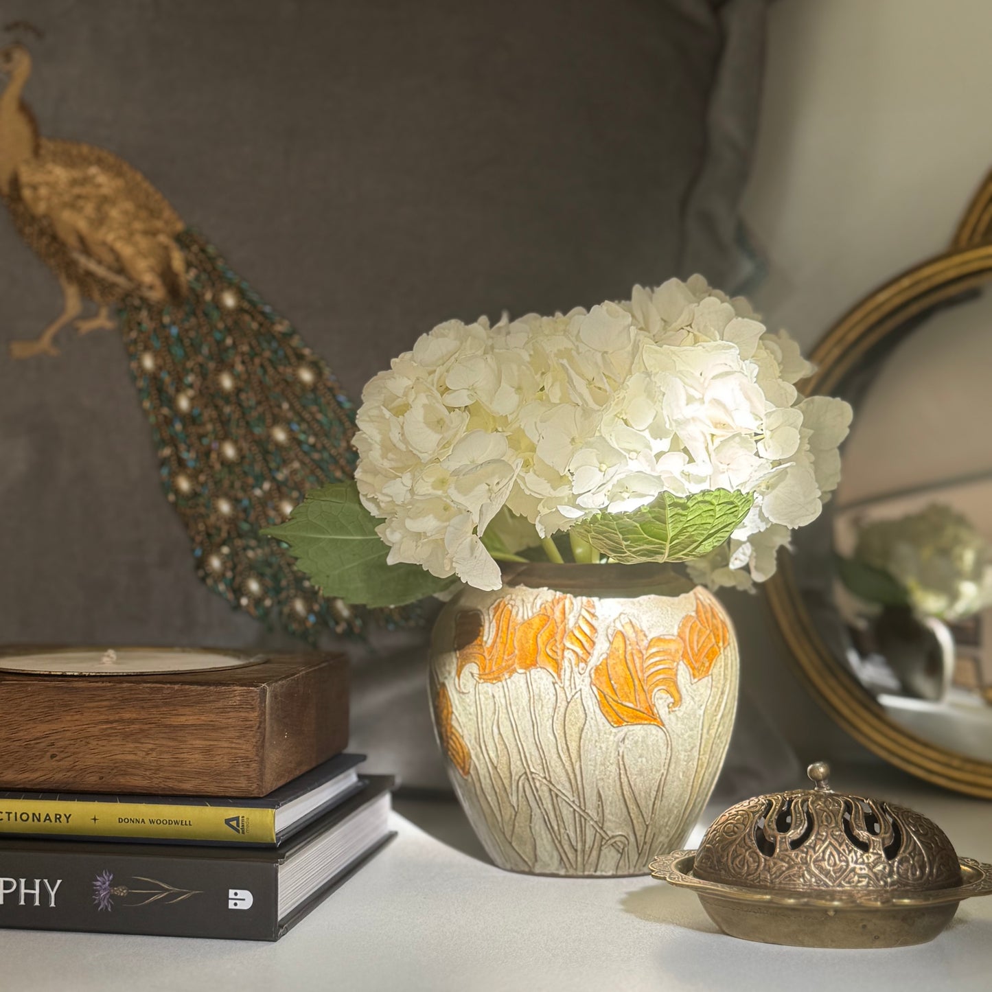 A brass and enamel iris vase with white hydrangea flowers sits on a shelf with decor items, books, and mirrors.