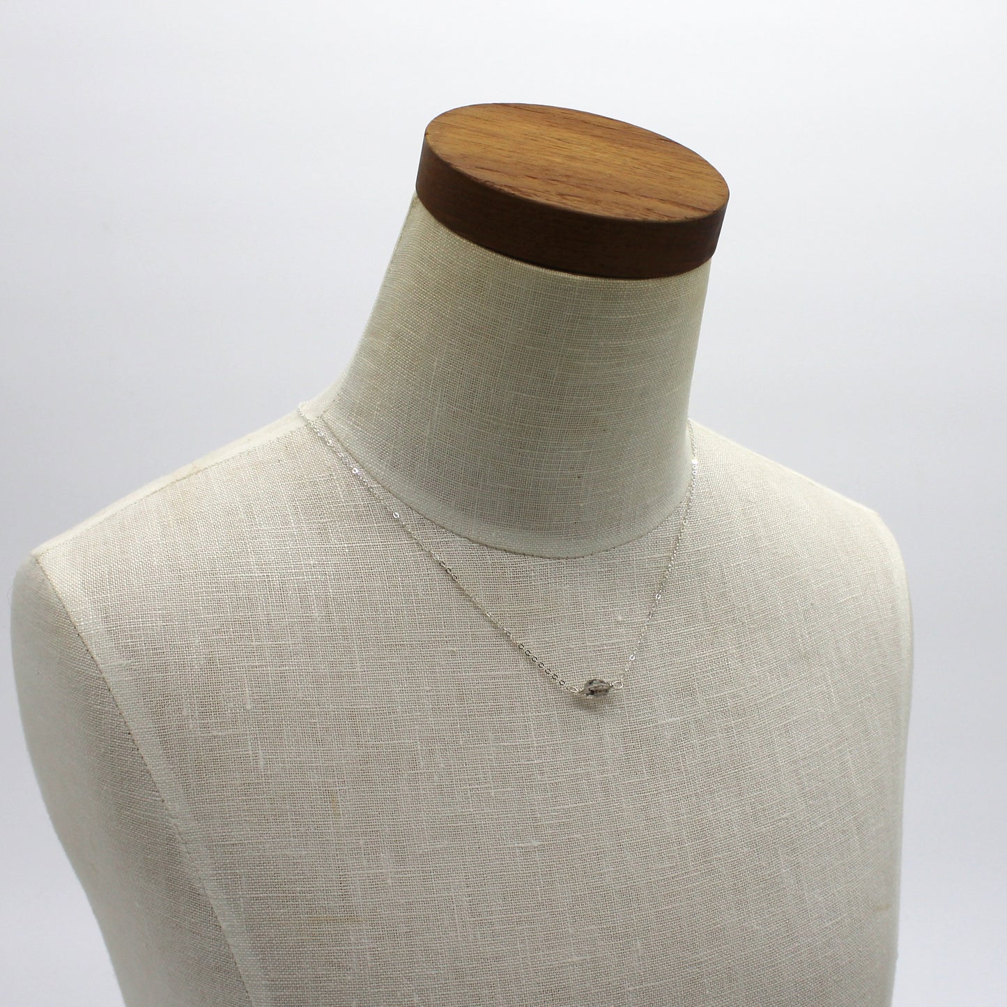 A dainty silver chain with a Herkimer diamond pendant on an ivory linen bust with a wooden neck.