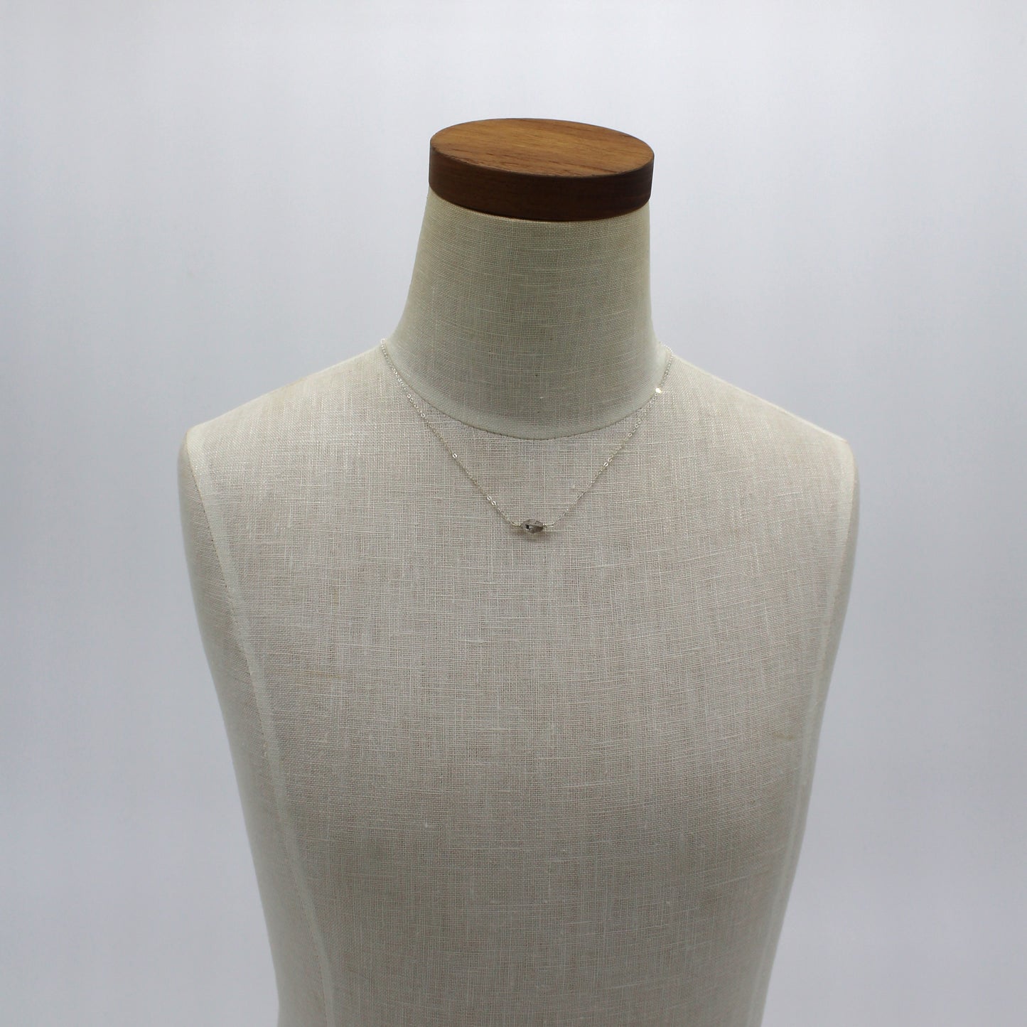 A dainty sterling silver chain featuring a Herkimer Diamond on an ivory linen bust with a dark wooden neck.