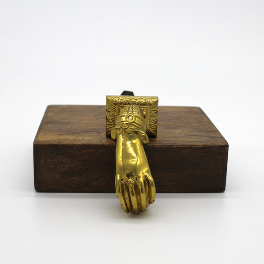 Front view of a polished brass door knocker in the shape of a ringed hand with sleeve at wrist sits on a wooden block with a white background.