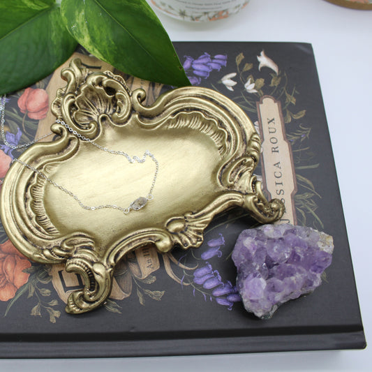 A sterling silver necklace featuring a small faceted Herkimer Diamond rests in a vintage golden jewelry tray sitting on a floral book with an amethyst cluster and leaves.