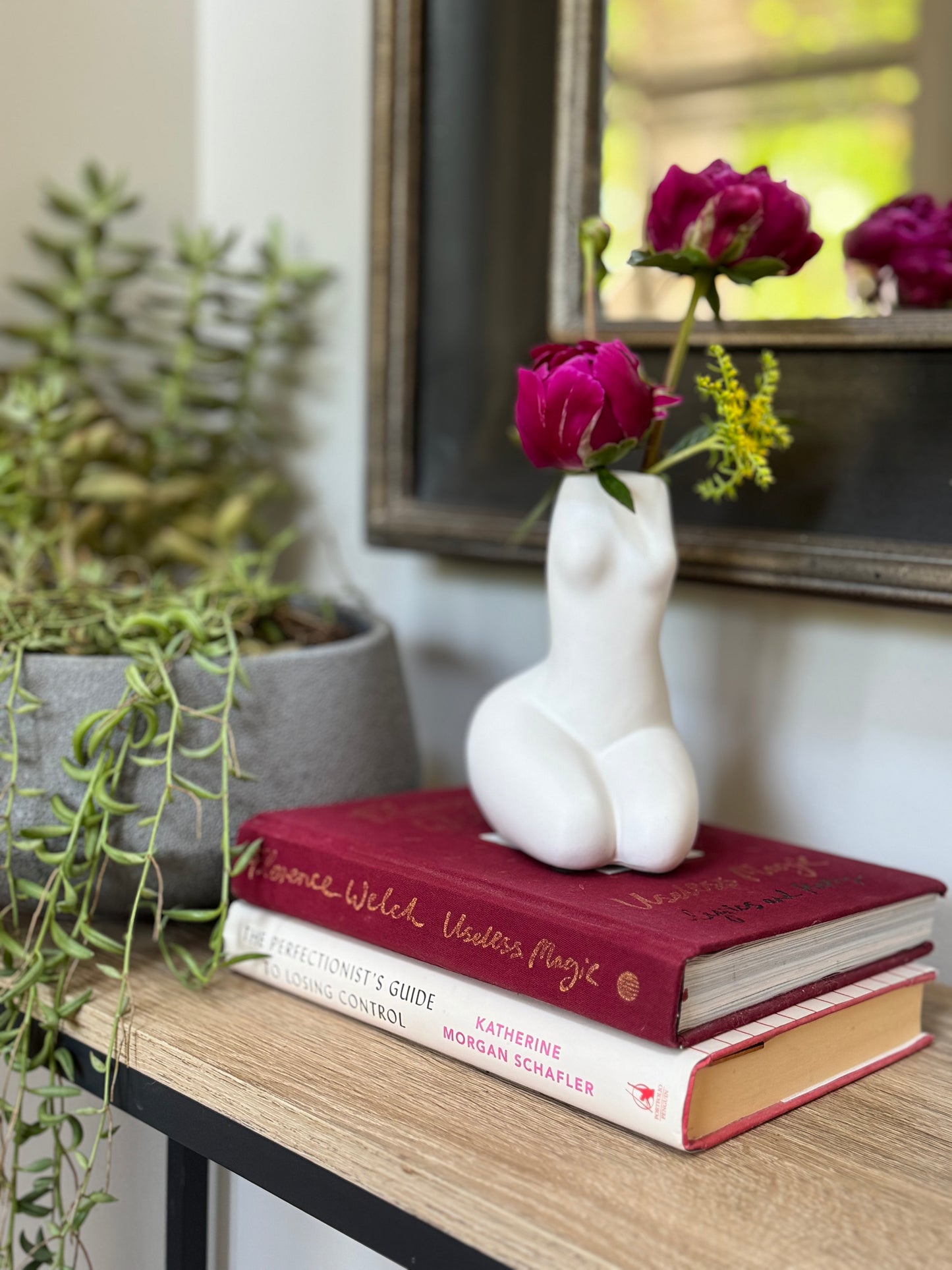 White ceramic sculpture of a nude figure with pink flowers on top of books, surrounded by plants and a mirror.