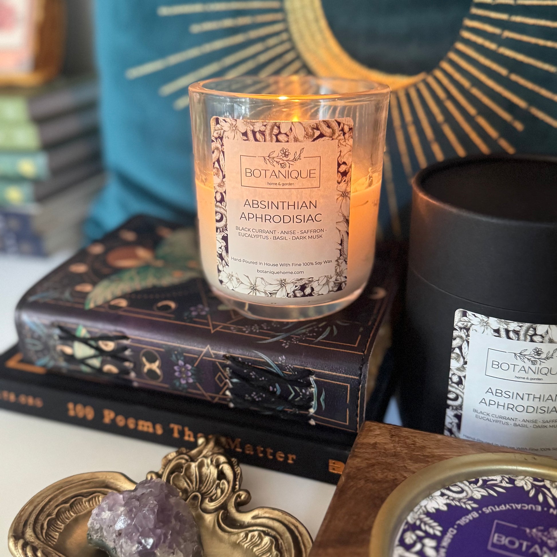 Candle labeled 'Botanique Absinthian Aphrodisiac' on books with decorative items in the background