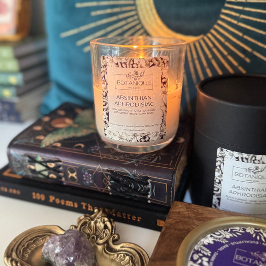 Candle labeled 'Botanique Absinthian Aphrodisiac' on books with decorative items in the background