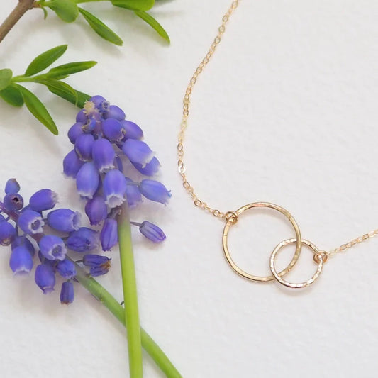 Gold necklace with interlocking rings next to purple flowers on a white background
