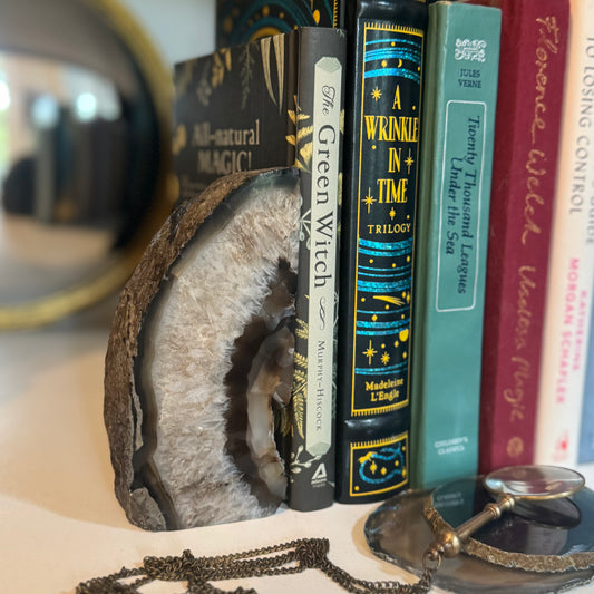 Geode Bookend holding up stack of books  on shelf with agate slices and vintage magnifying glass in front and round mirror behind.