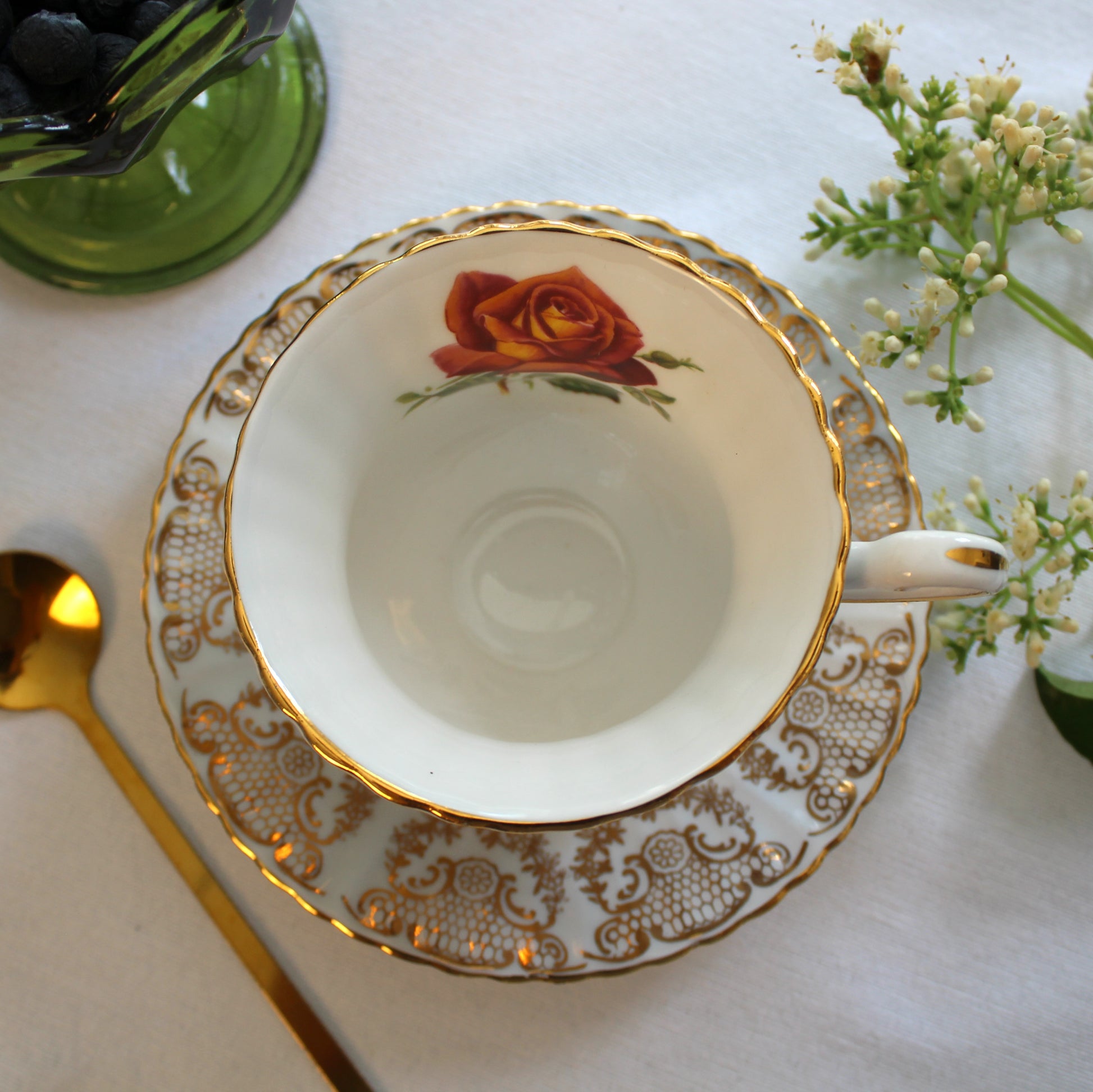 Teacup with floral design on a white tablecloth with a gold spoon and green vase.