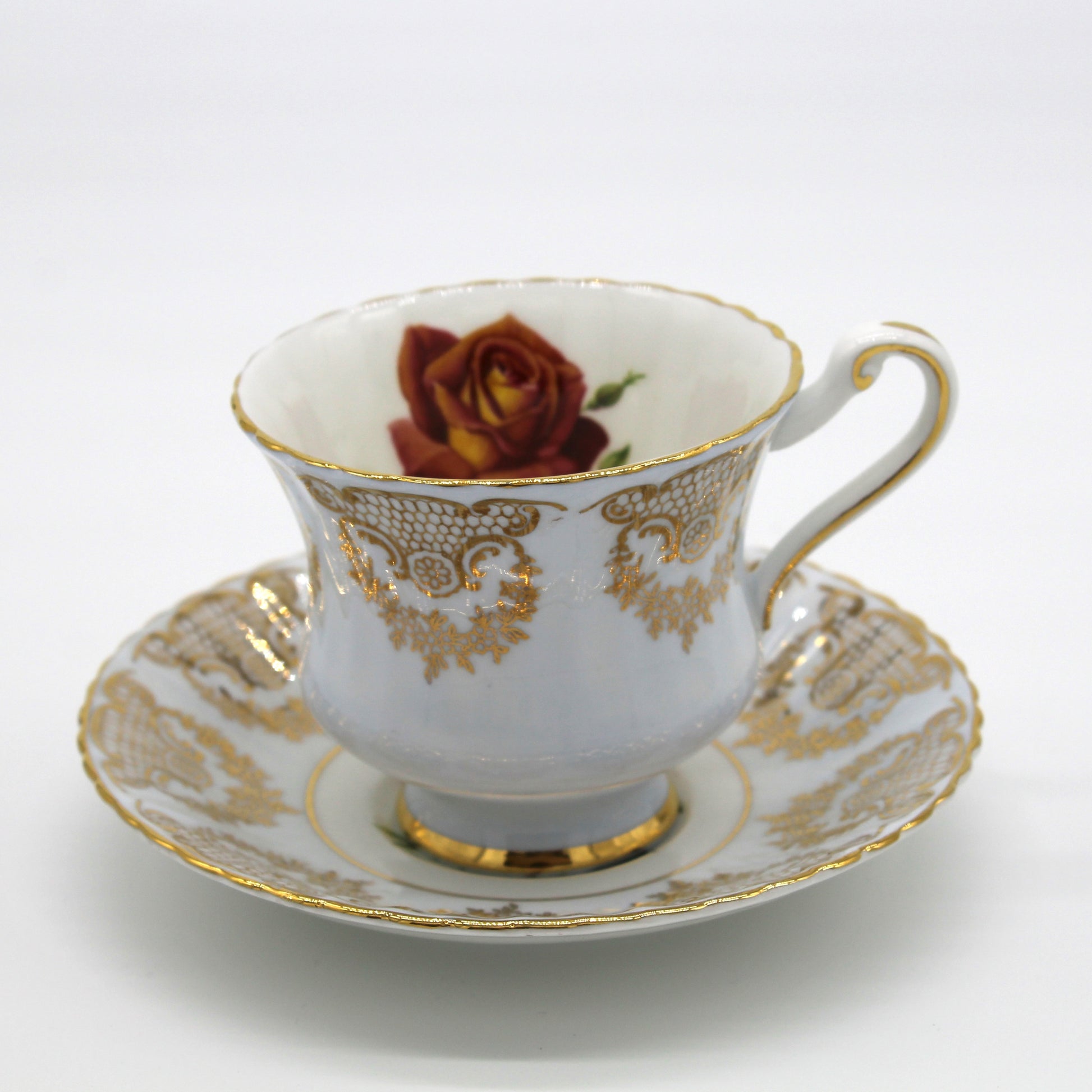 White teacup with gold detailing and a red rose on a matching saucer against a white background